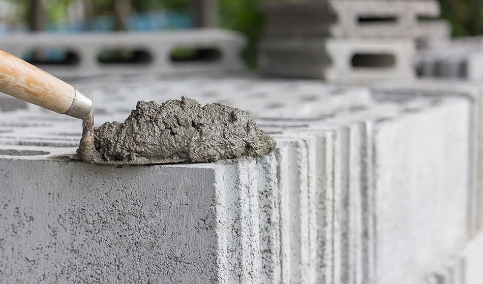 The image shows a close-up of a masonry trowel spreading wet cement mortar onto the top edge of a concrete block. The concrete blocks are stacked, with visible hollow cores, typical in construction for building walls. The fresh mortar appears thick and textured, ready to bond the blocks in place.