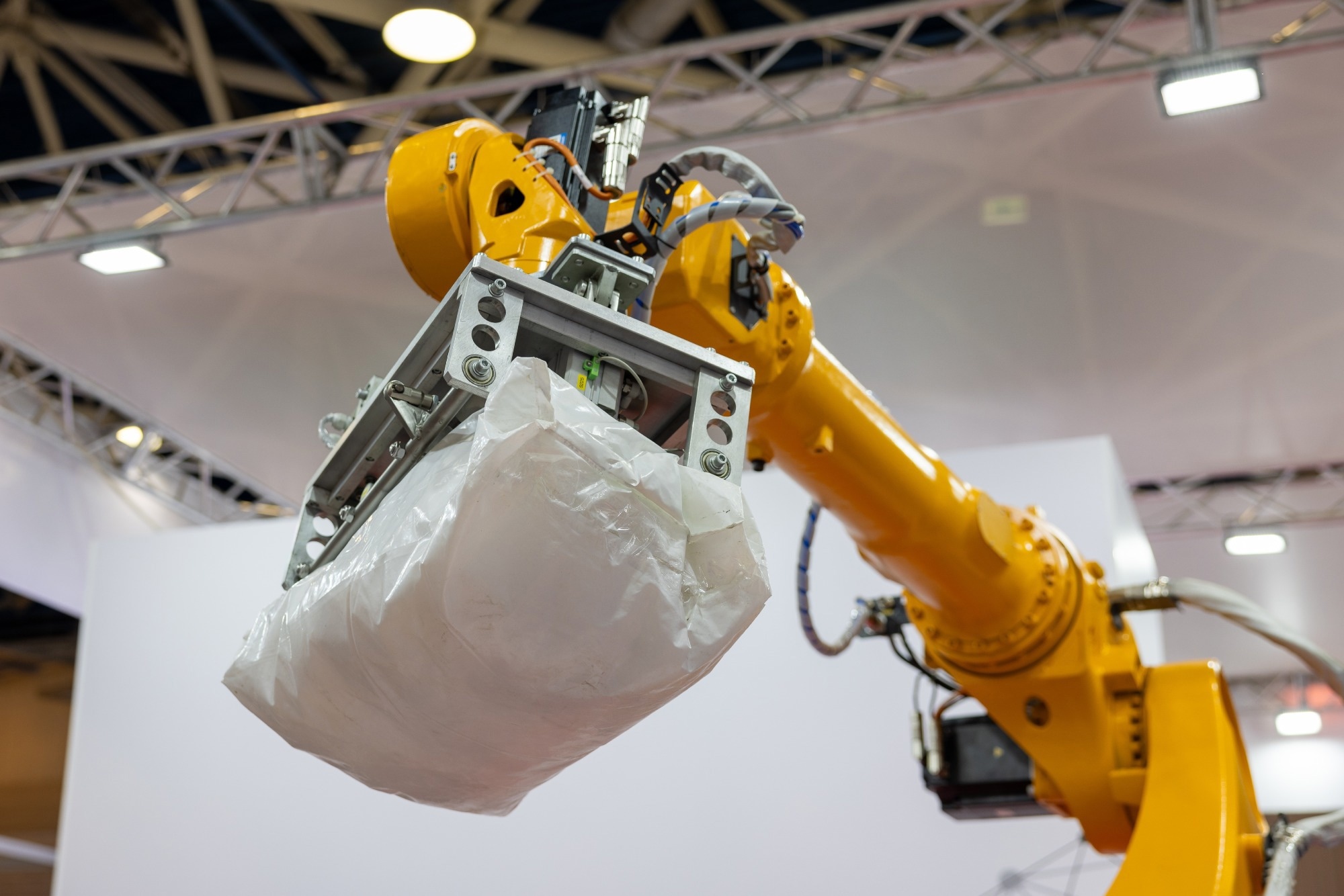 An industrial robotic arm with a pneumatic gripper lifts a heavy white bag in a factory setting.