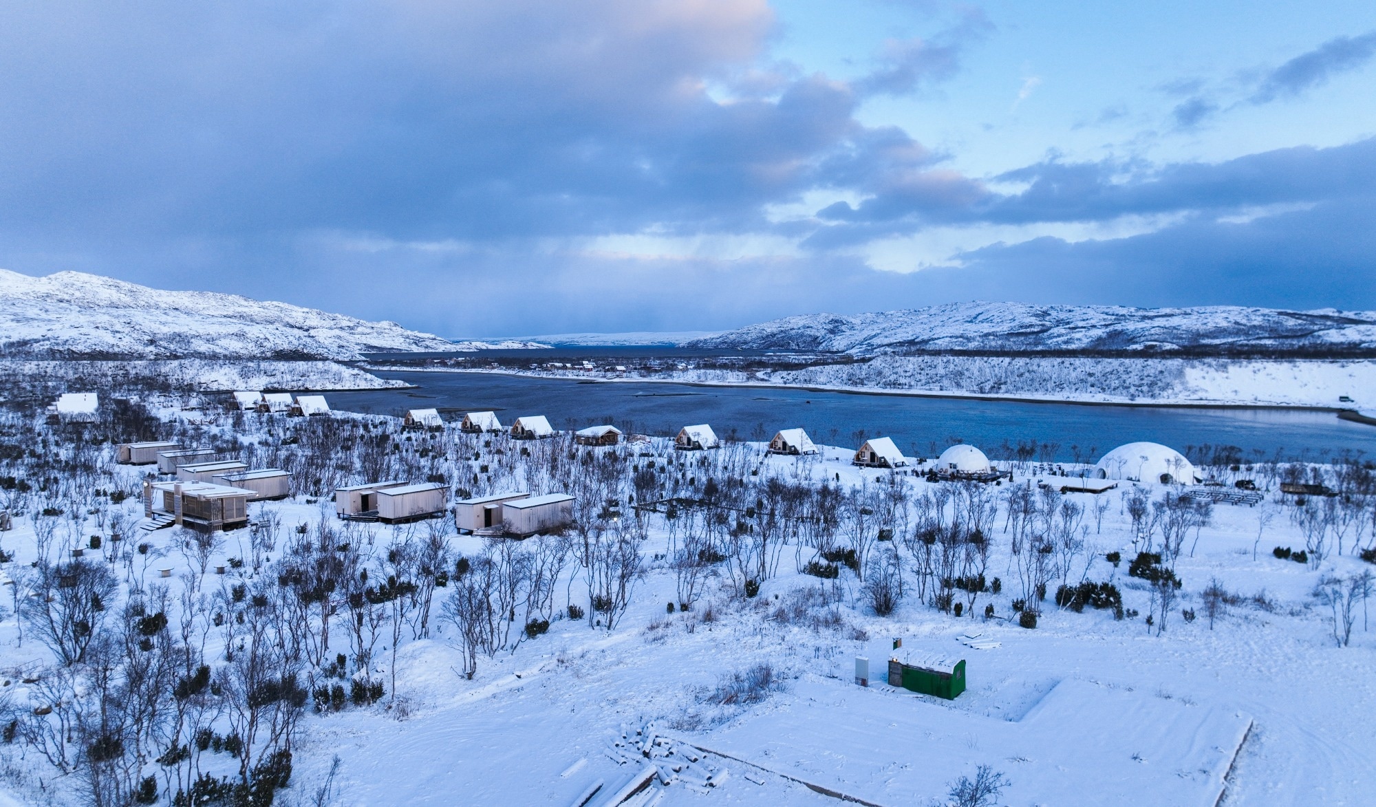 Aerial view of an Arctic tourist camp featuring modern wooden cabins and a white dome structure near a partially frozen body of water, surrounded by snowy hills