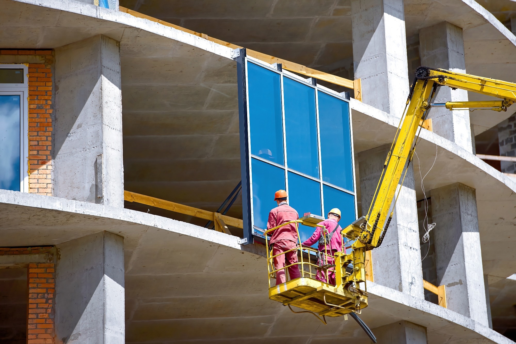 Workers in lift bucket installing large glass panes on new building at construction site.