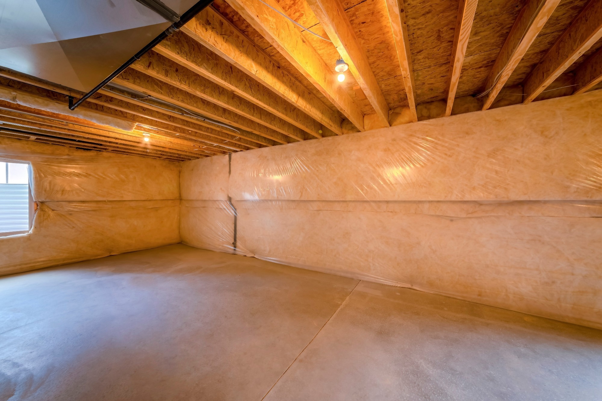 Unfinished basement with wall insulator and plastic vapor barrier under the timber ceiling. There is a window on the left allowing the natural light to enter the basement with concrete flooring.