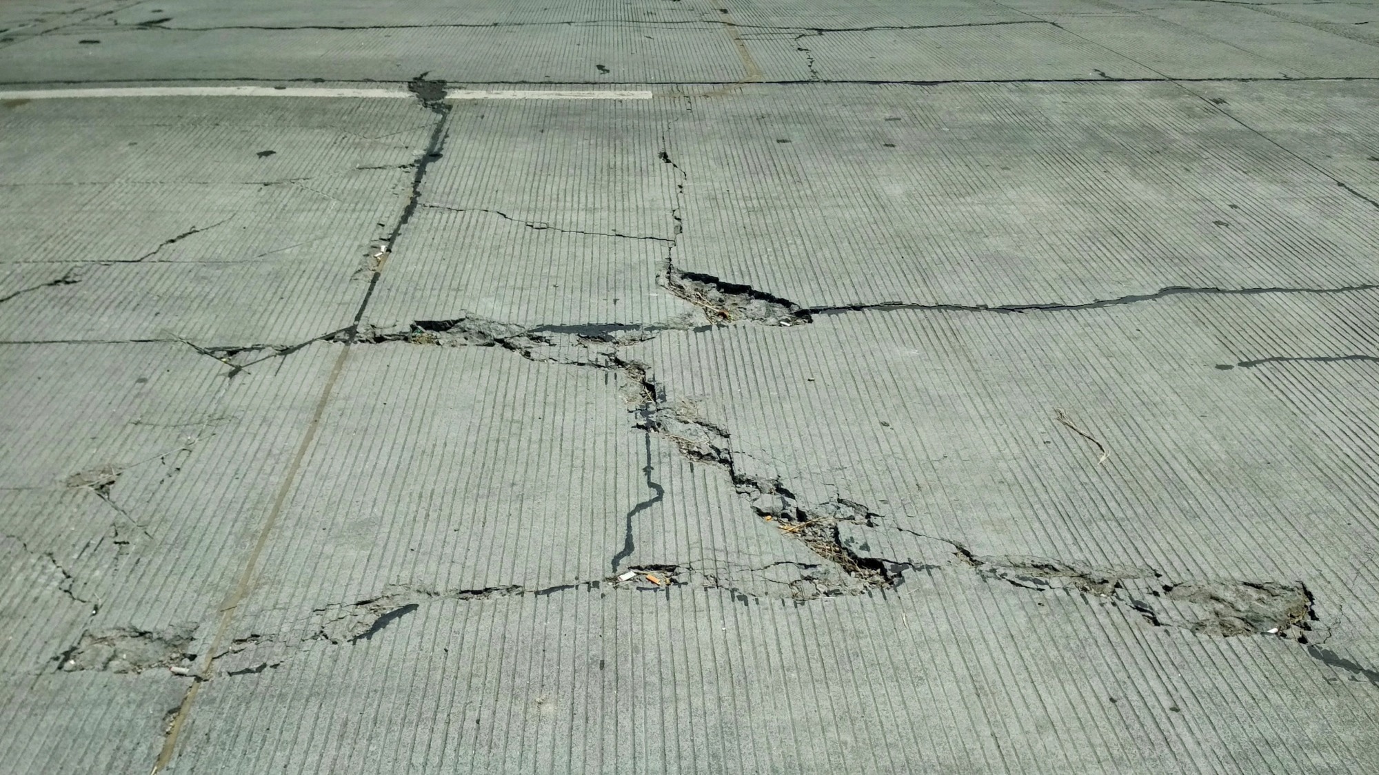 Heavily cracked concrete road surface with visible fractures and signs of deterioration.