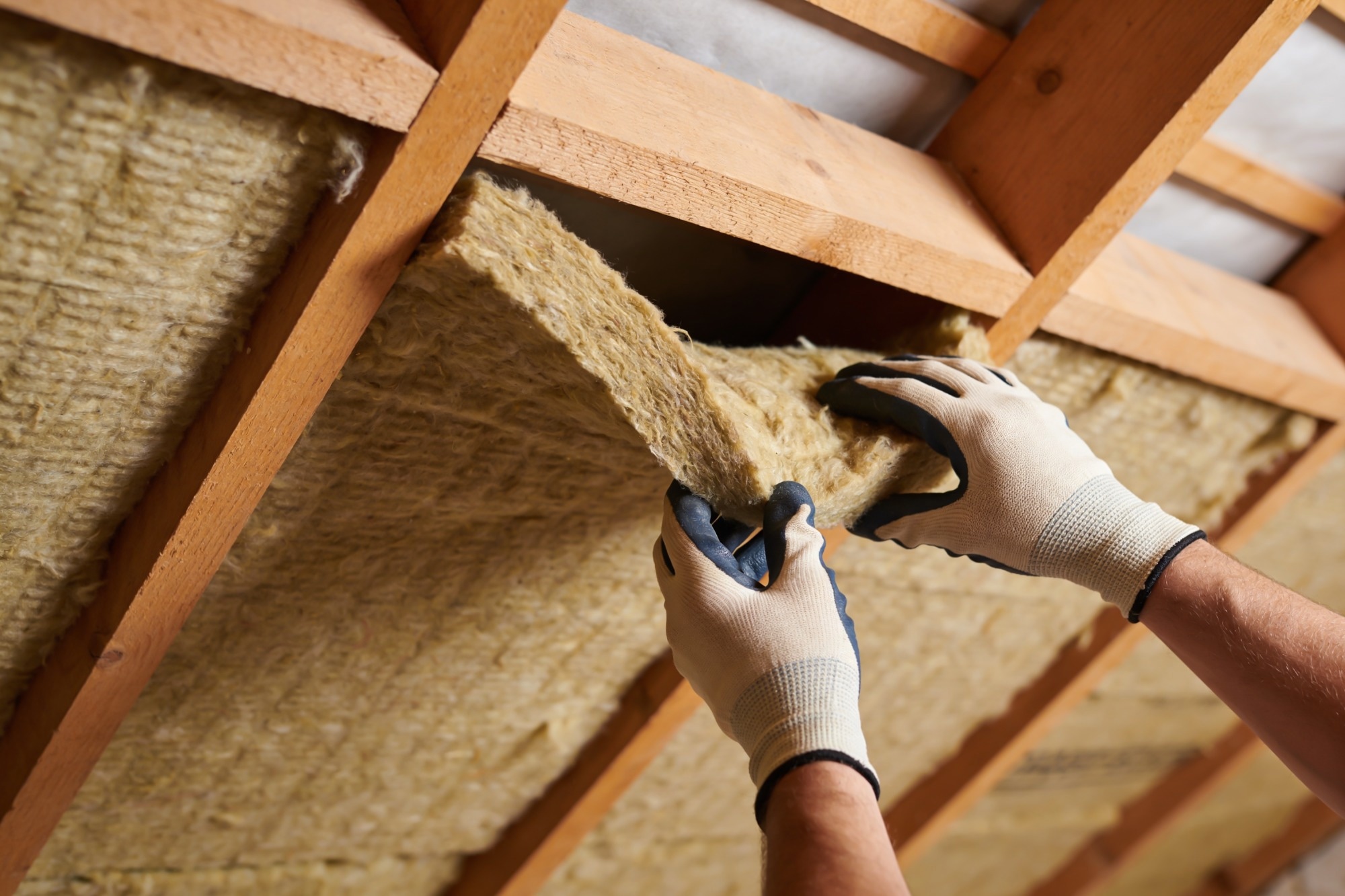 Close-up of hands wearing gloves carefully placing mineral wool insulation between wooden beams in a building attic.