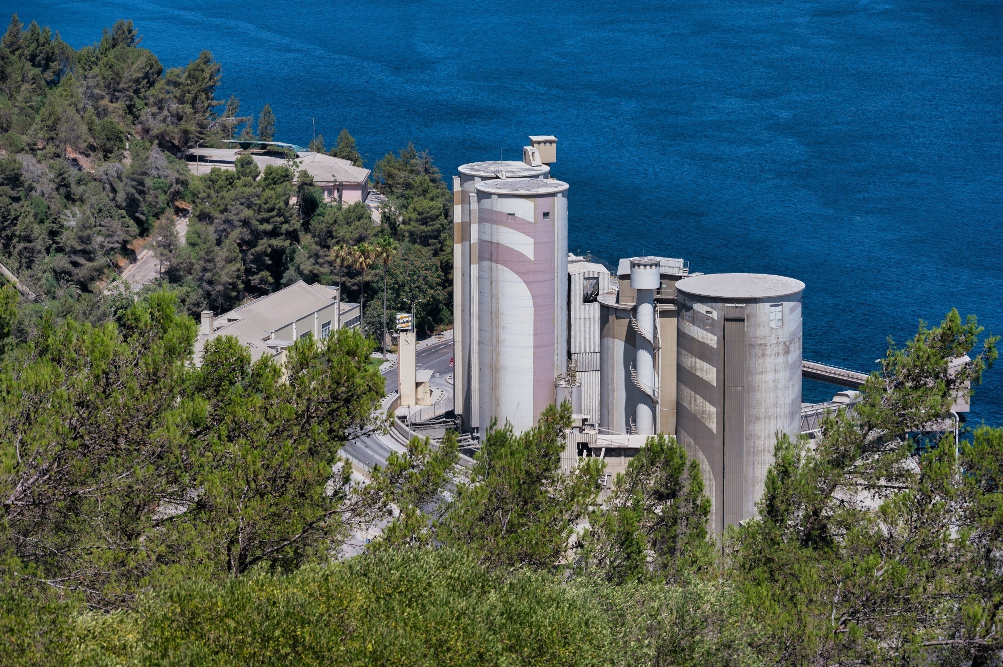 Cement plant with large silos located on the coast within the protected Arrábida Natural Park in Portugal, surrounded by forested hills.